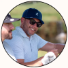 A man wearing a navy blue deer logo baseball cap, black sunglasses, and a light blue patterned polo shirt, smiling outdoors on a golf course. The photo is framed in a circular border with another person partially visible on the left.