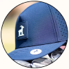 A close-up circular shot of a navy blue baseball cap with a black square patch featuring a white goat logo on the side. The cap has perforated side panels and a small white logo patch on the visor, with golf clubs partially visible underneath.